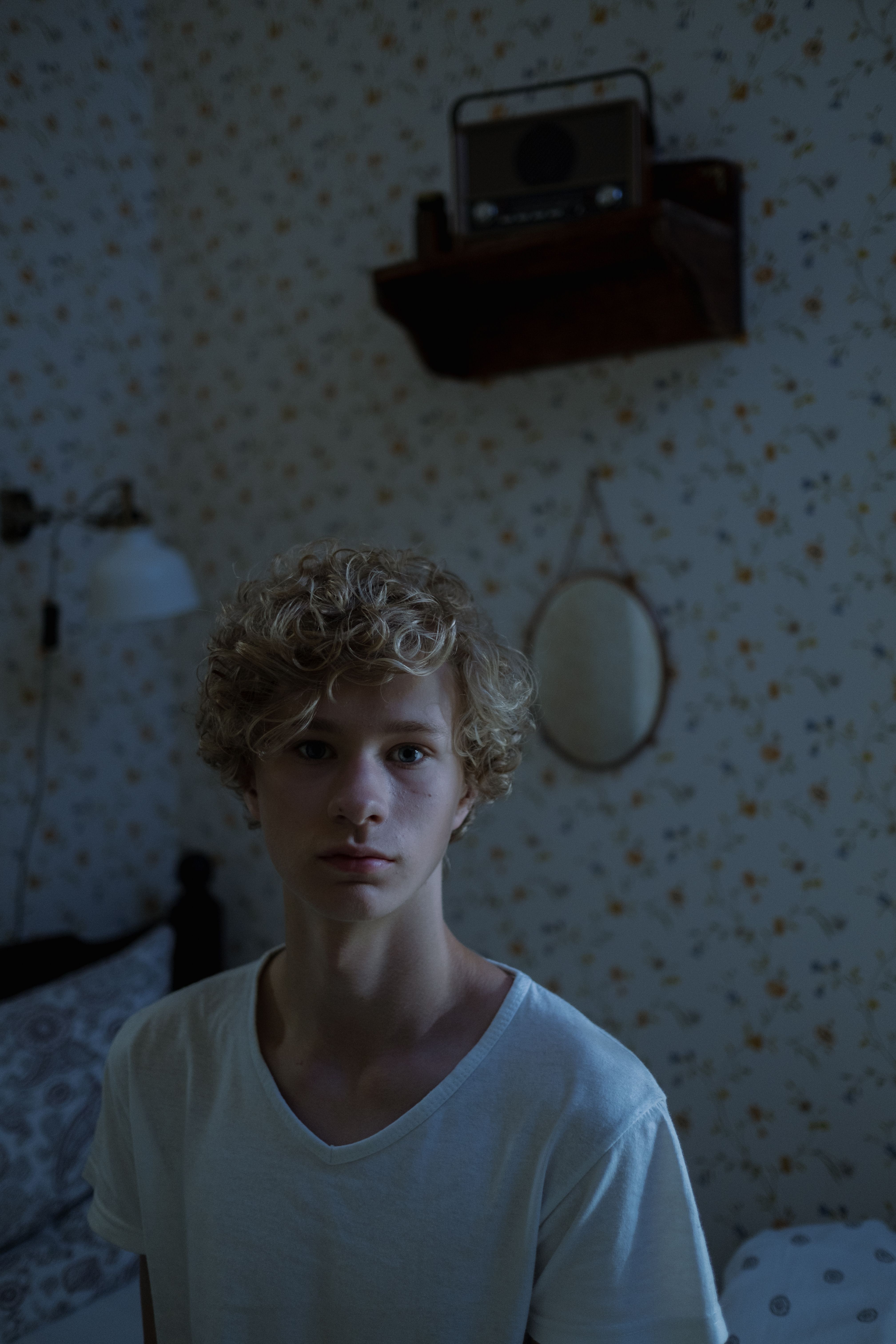 Teen boy in dimly lit vintage bedroom with radio and mirror on floral wallpaper.