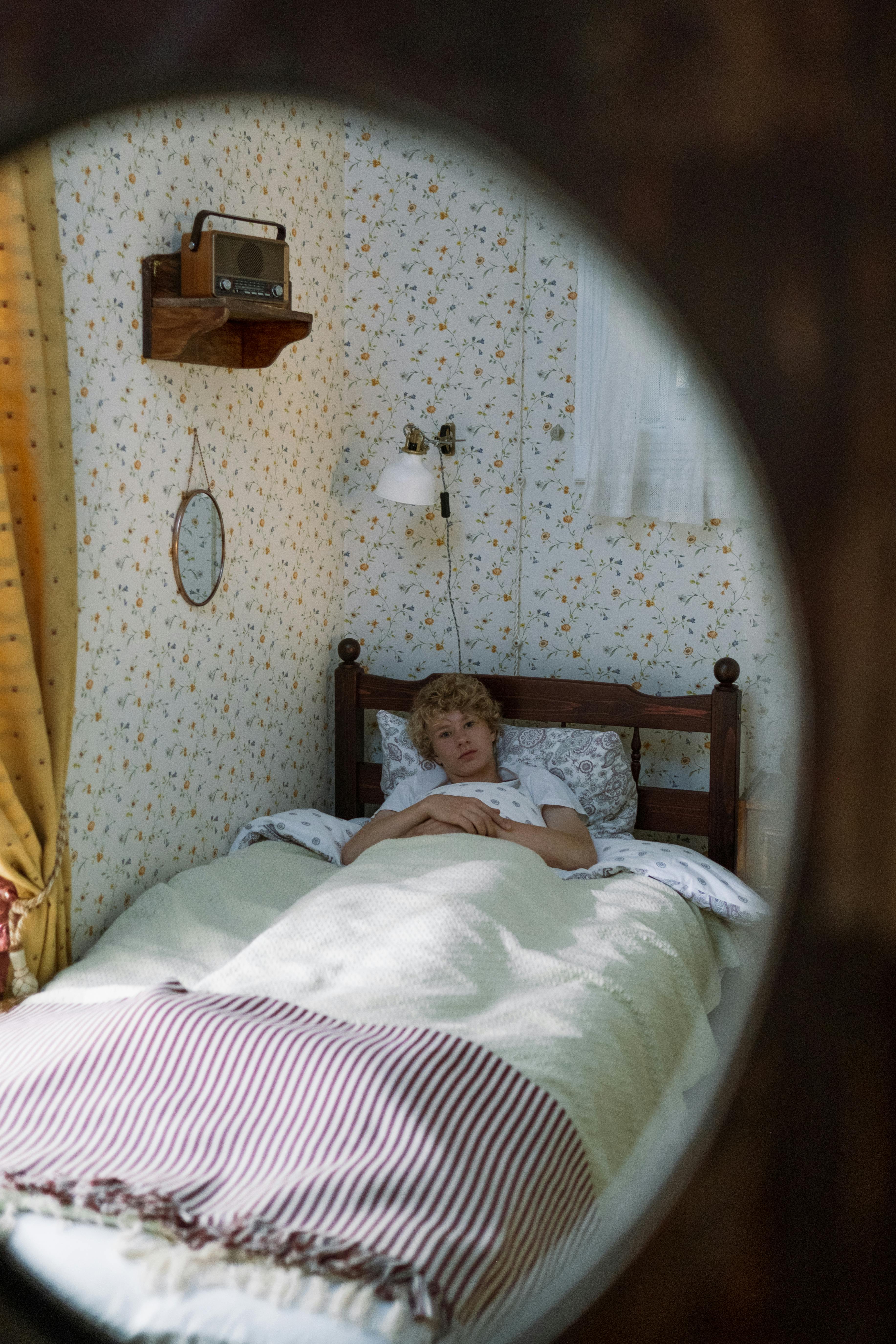 Teenage boy with curly hair relaxing in a cozy vintage-style bedroom reflected in a mirror.