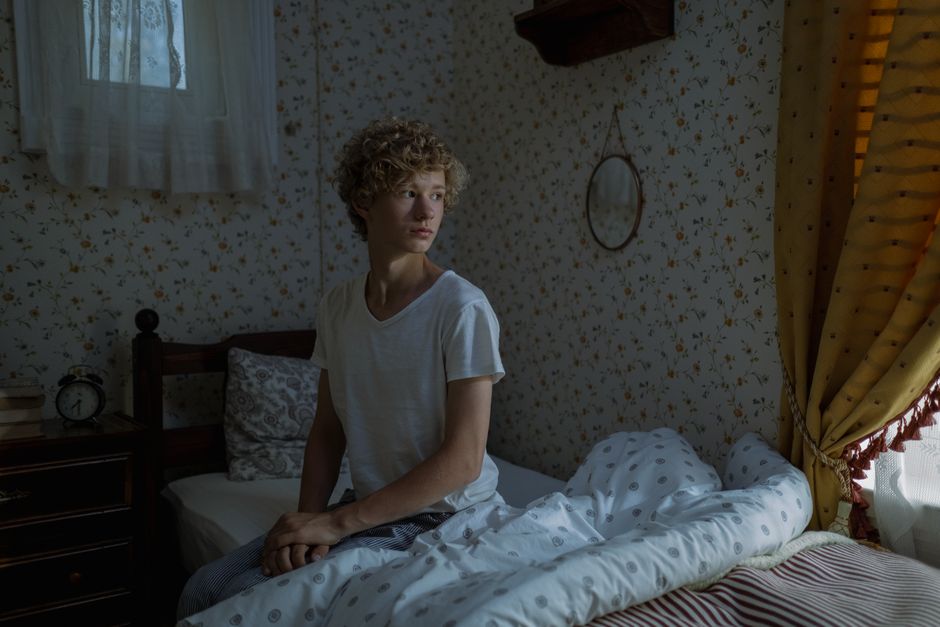 Teenage boy with curly hair sitting on a bed in a cozy, dimly lit bedroom.