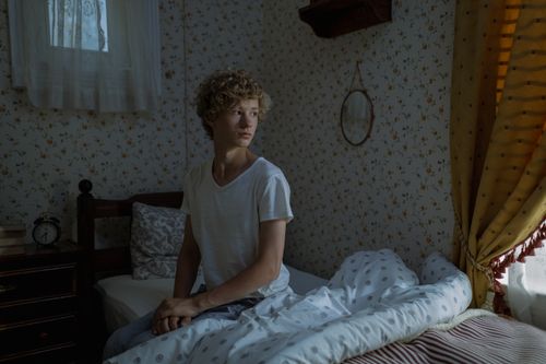 Free Teenage boy with curly hair sitting on a bed in a cozy, dimly lit bedroom. Stock Photo