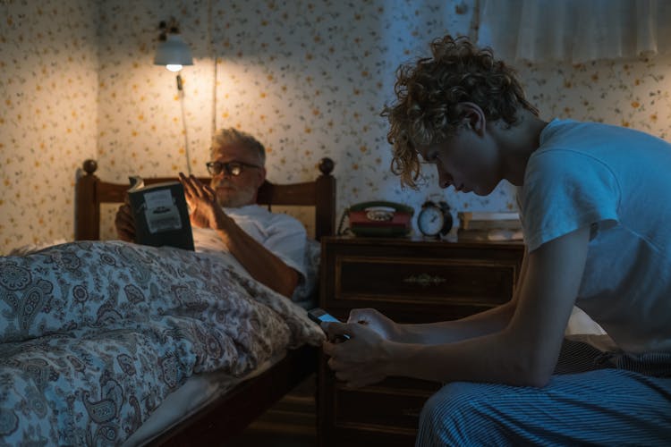 Man Reading In Bed And Boy Using Smart Phone 