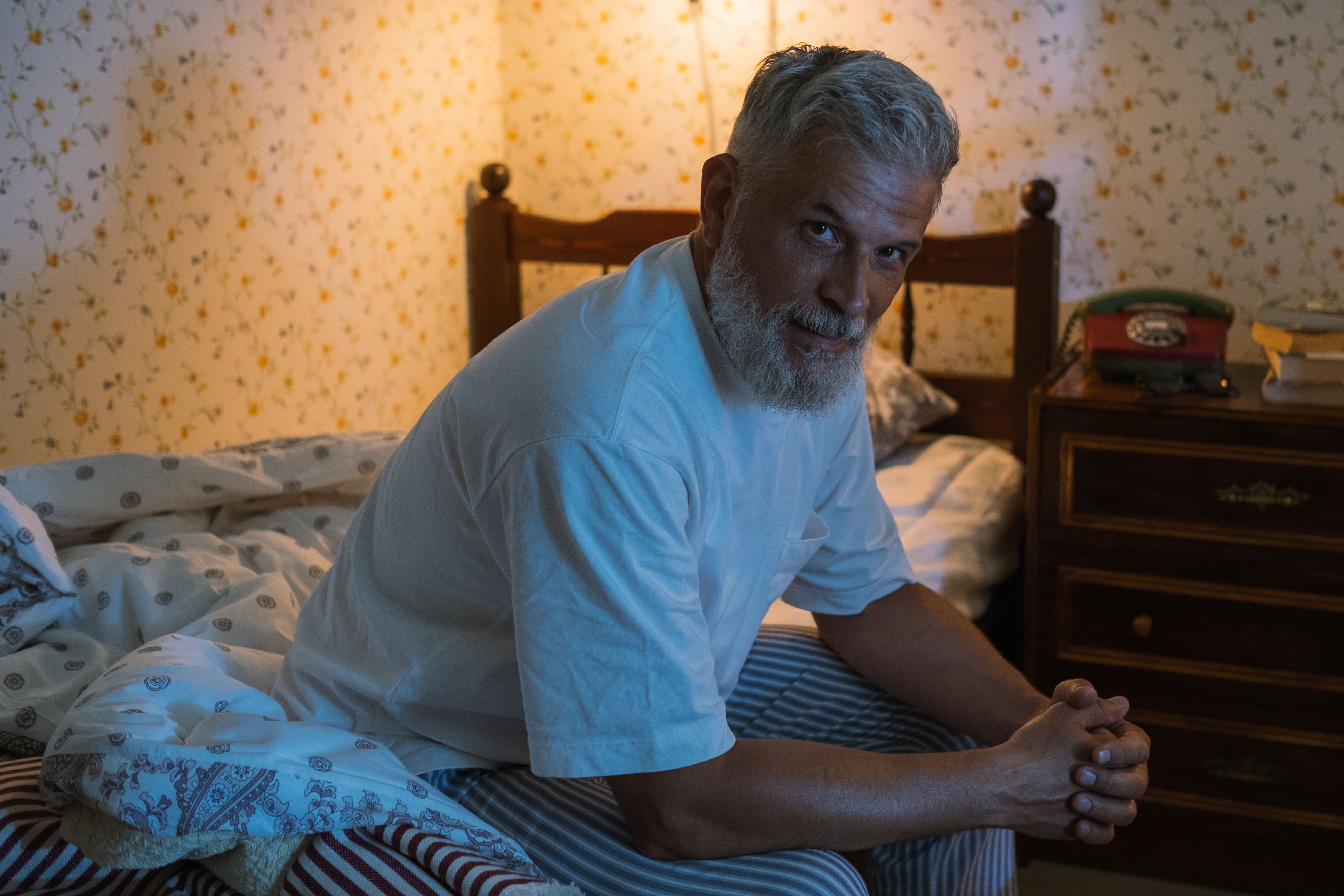 Older man sitting on the edge of his bed at night, looking tired