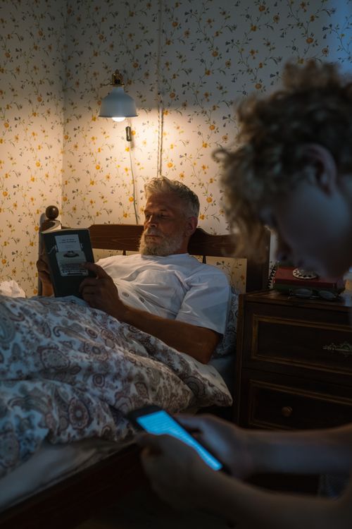Free Elderly man reading in bed while grandson uses smartphone. Stock Photo