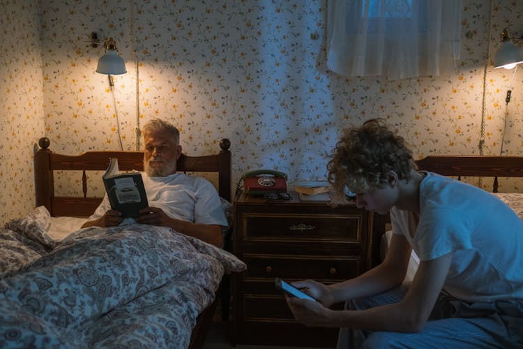 Man Reading In Bed And Boy Using Smart Phone 