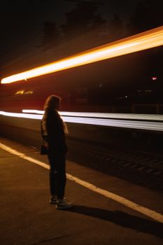 A woman stands on a platform as a train speeds by, creating light trails during nighttime.