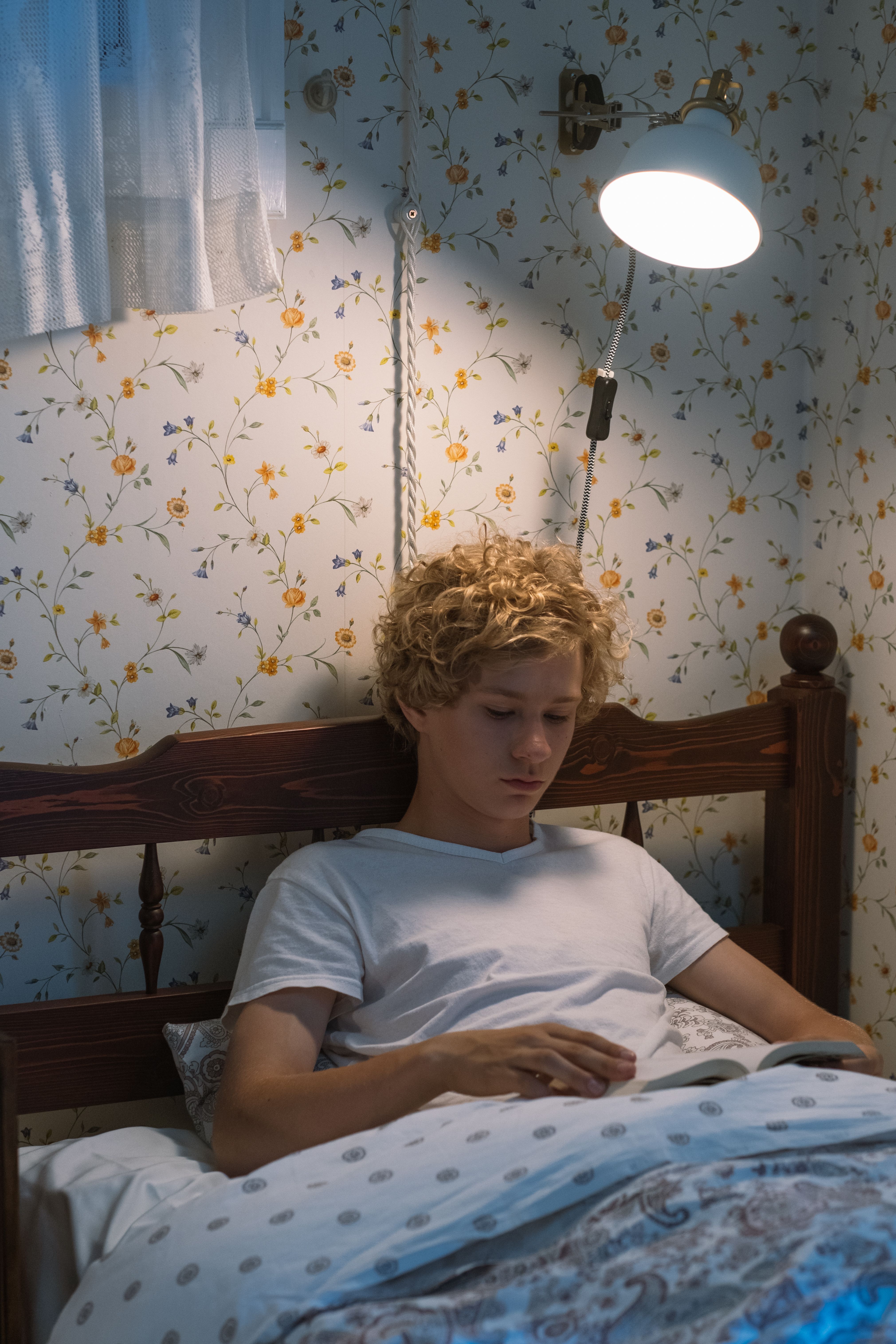 A teenager lies in bed, reading under a lamp in a cozy, floral-wallpapered room.