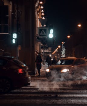 A moody nighttime cityscape with cars, passersby, and illuminated streetlights casting a warm glow.