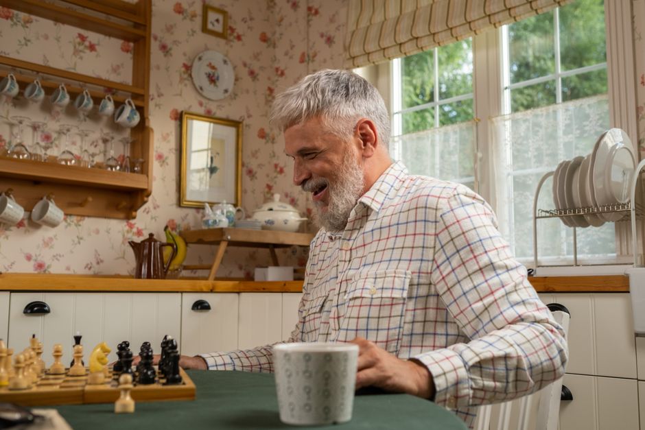 Smiling elderly man in a plaid shirt playing chess in a warm, floral kitchen setting, feeling relaxed.