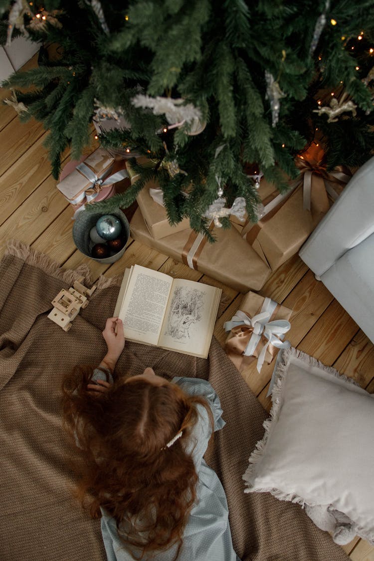 Teenage Girl Laying On Floor By Christmas Tree And Reading Book