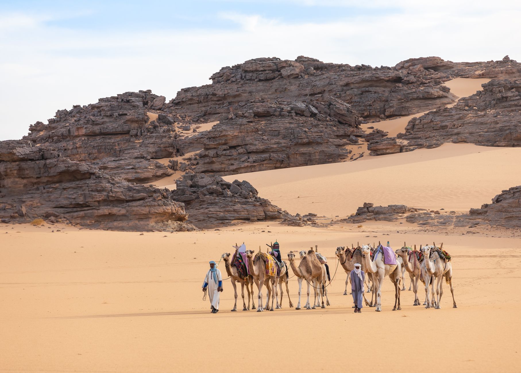 Camels and people traverse rocky desert terrain in Illizi, Algeria.