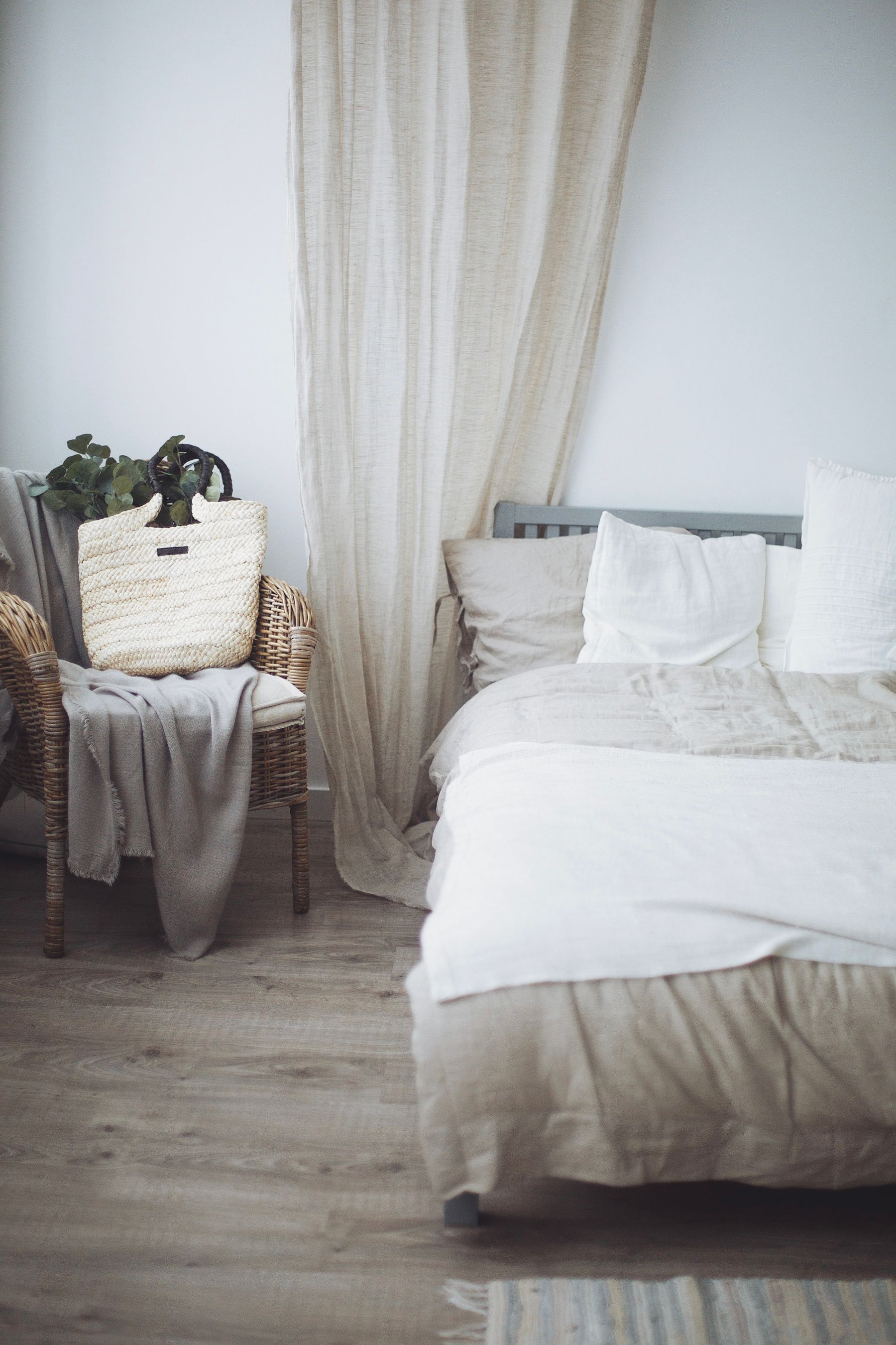 Bedroom corner with a woven rattan chair, pale wall, wood floor and soft neutral textiles