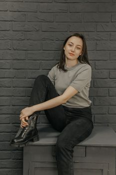 Stylish woman in gray shirt sitting against brick wall with black pants and boots.