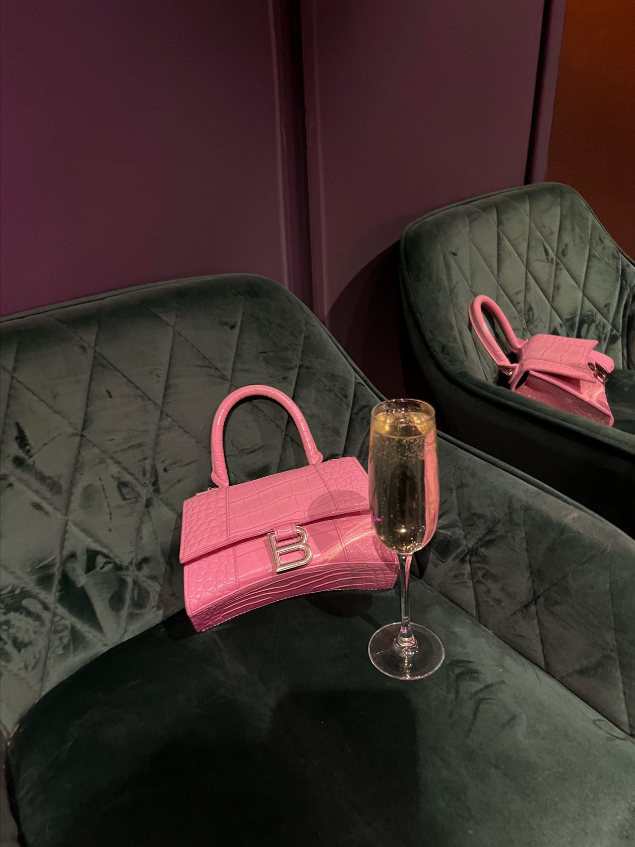Pink croc-embossed handbag and a glass of champagne on green velvet chairs
