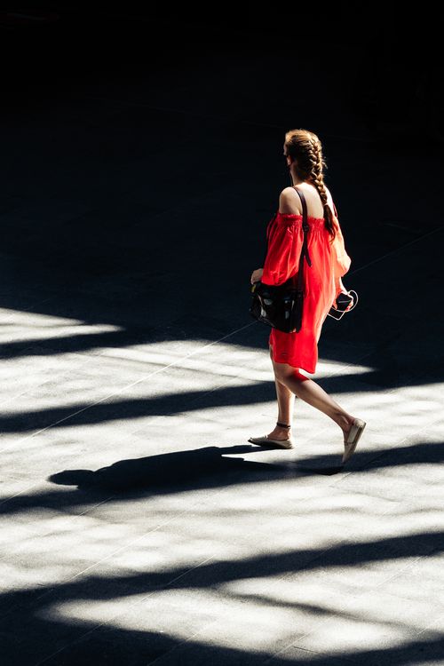 Free A woman in a red dress walks across a sunlit city pavement, casting a shadow. Stock Photo