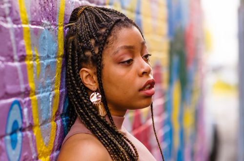 Free A woman with braided hair leans against a colorful mural wall outdoors, deep in thought. Stock Photo