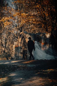 A newlywed couple walking under a stone archway amidst autumn leaves.