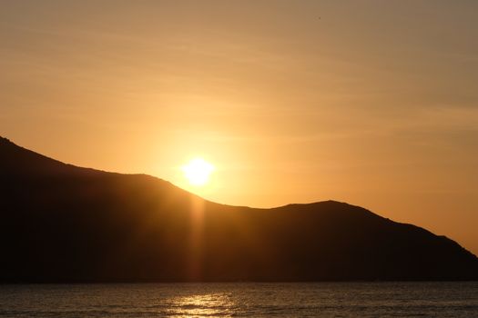 A tranquil sunset over the ocean with silhouetted mountains and a glowing sun.