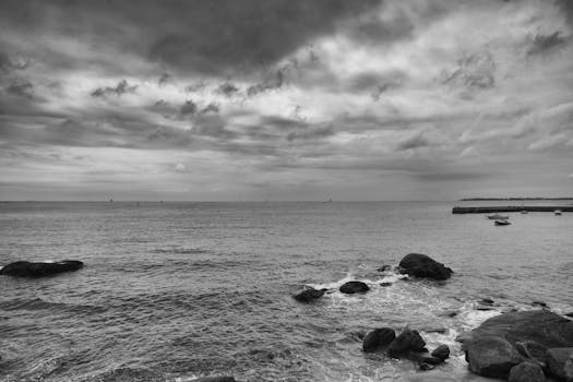 Black and white ocean view under a dramatic cloudy sky, showcasing rocks in the water.