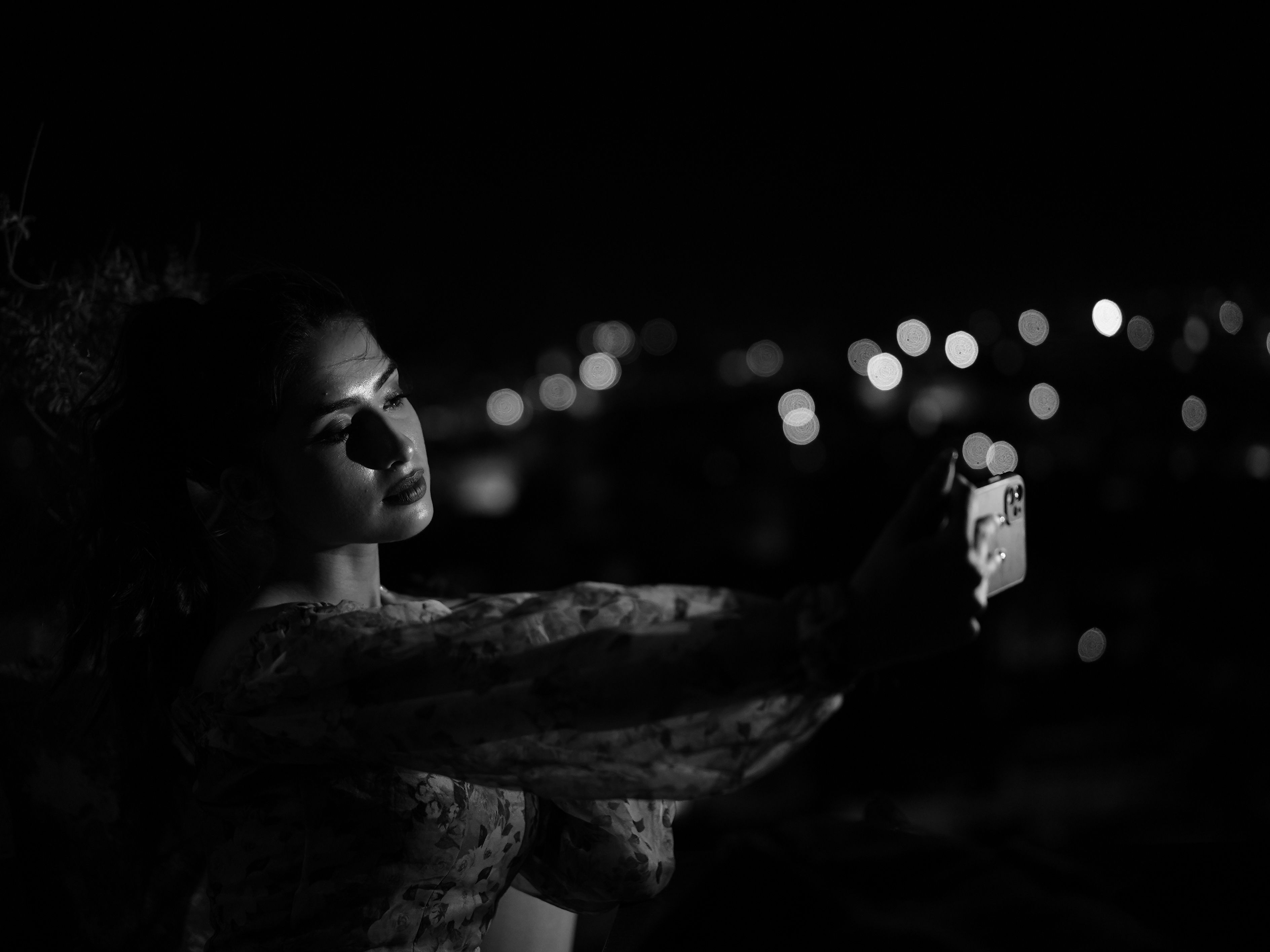 A woman takes a monochrome selfie at night, capturing the dramatic city lights surrounding her.