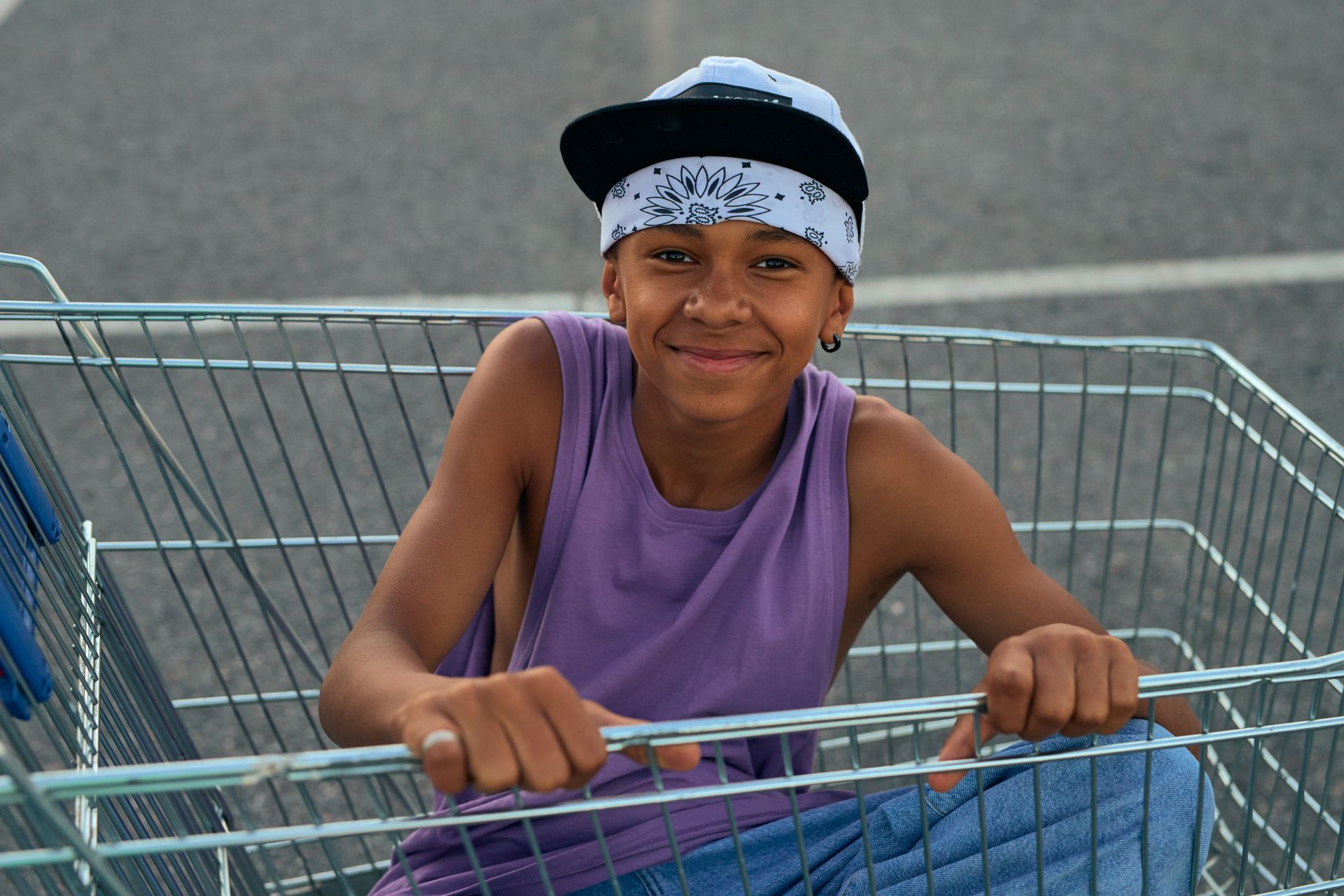 Smiling boy sits in a shopping cart outdoors wearing a cap and bandana.