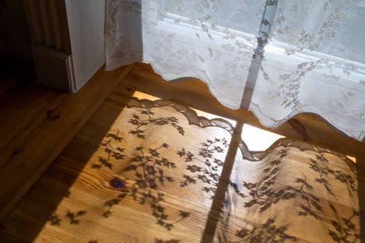 Lace curtains casting delicate floral shadows on a hardwood floor in warm sunlight, creating a vintage ambiance.
