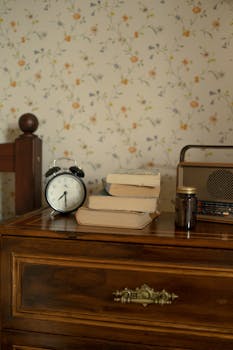 A cozy vintage bedroom setup featuring an alarm clock, books, and retro radio on a wooden chest of drawers.