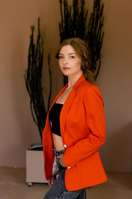Woman in vibrant orange jacket poses elegantly indoors with a cactus backdrop.