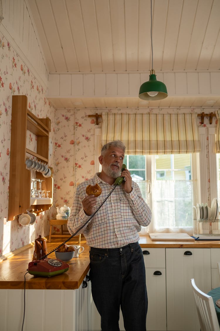 Senior Man Talking On Landline Phone And Eating Cinnamon Bun In Old-Fashioned Kitchen