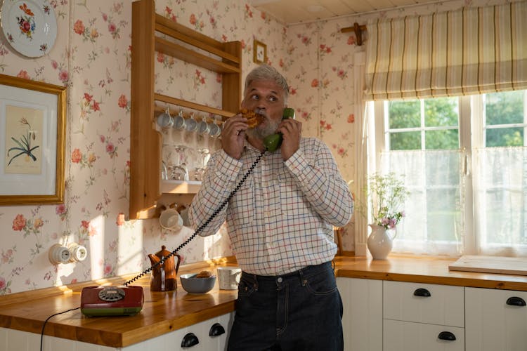 Senior Man Talking On Landline Phone And Eating Cinnamon Bun In Old-Fashioned Kitchen