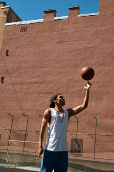 Man in a white tank top skillfully spins basketball on finger against urban backdrop.