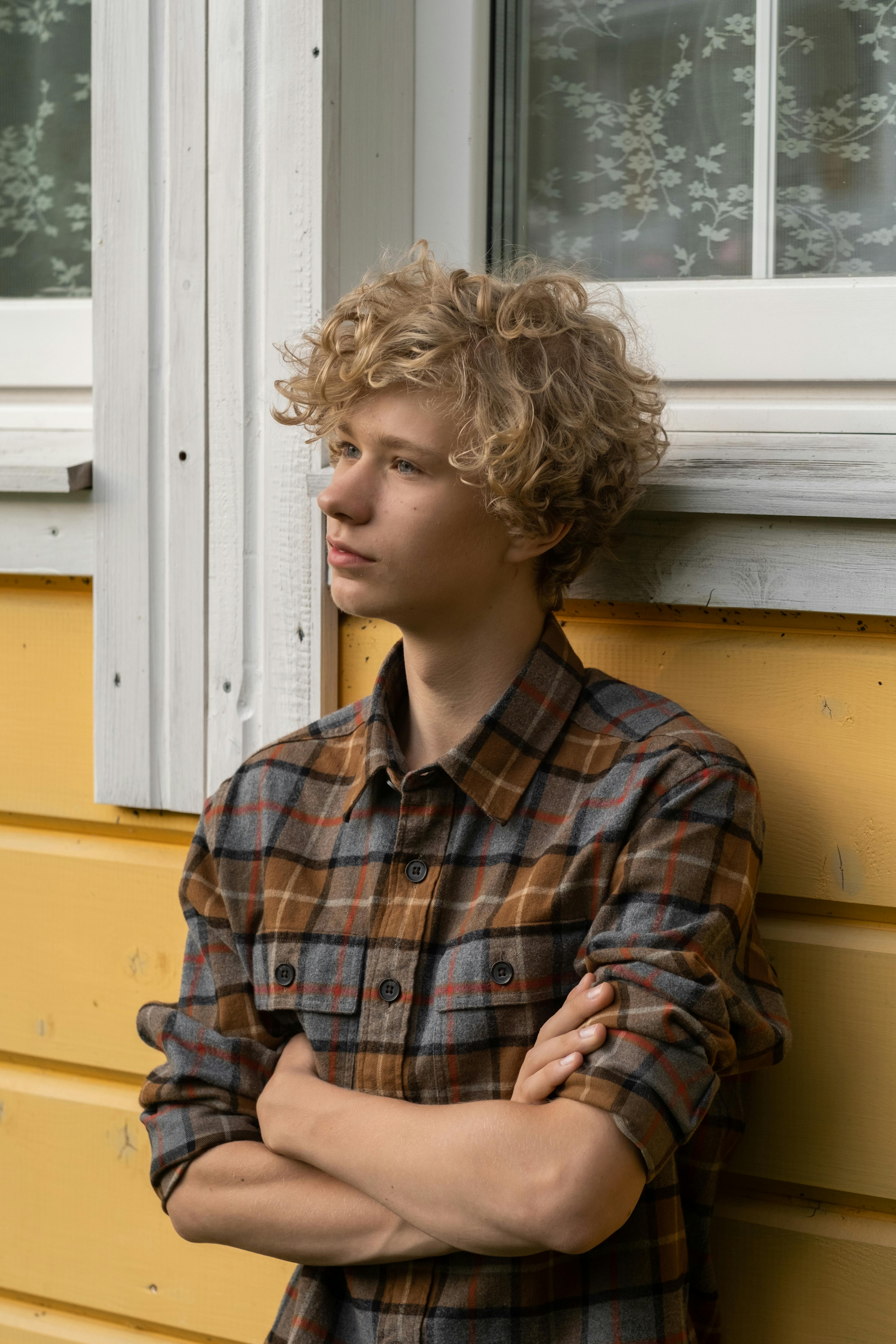 Teenage boy with curly blond hair and a plaid shirt standing with arms crossed by a yellow wall and window.