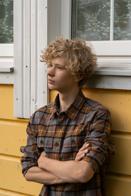 Free Teenage boy with curly blond hair and a plaid shirt standing with arms crossed by a yellow wall and window. Stock Photo