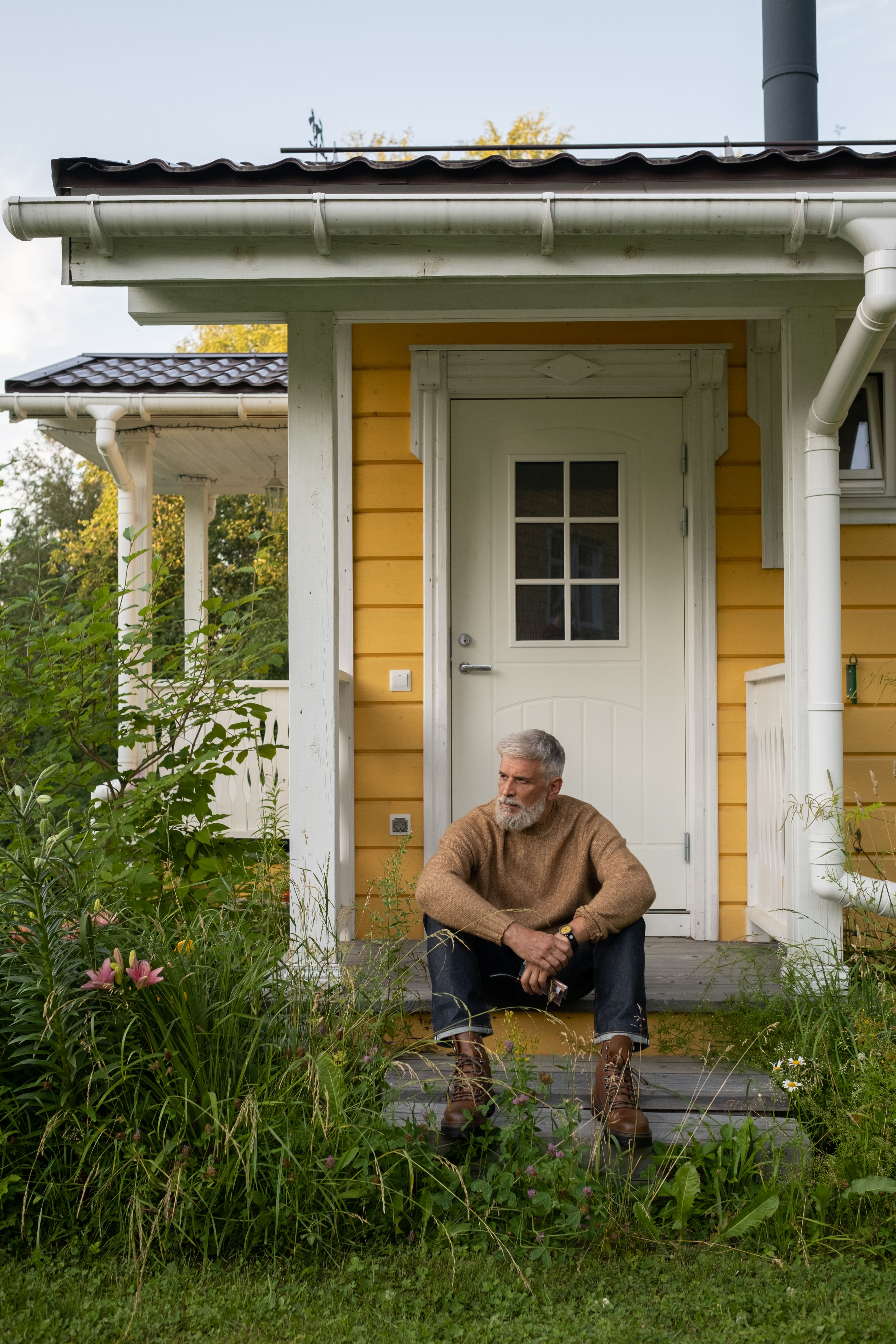 Senior man with gray beard sitting on porch steps of a quaint yellow house, surrounded by greenery.