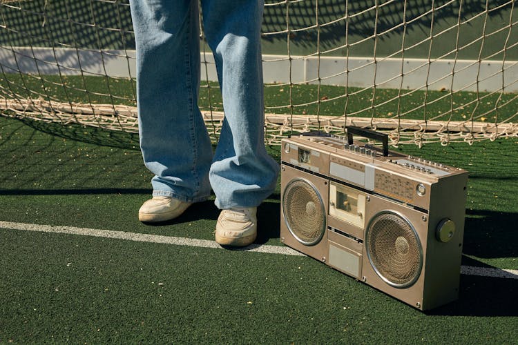 Person In Blue Denim Jeans And White Sneakers Standing Beside A Retro Boombox