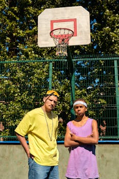 Two young men in vibrant attire pose confidently at an outdoor basketball court.