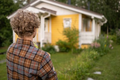 Free Teen with curly hair in plaid shirt stands facing a charming yellow house amidst lush greenery. Stock Photo