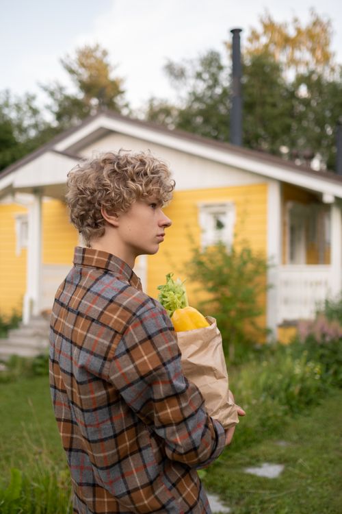 Free A teenager with curly hair stands outdoors holding groceries in front of a yellow house. Stock Photo