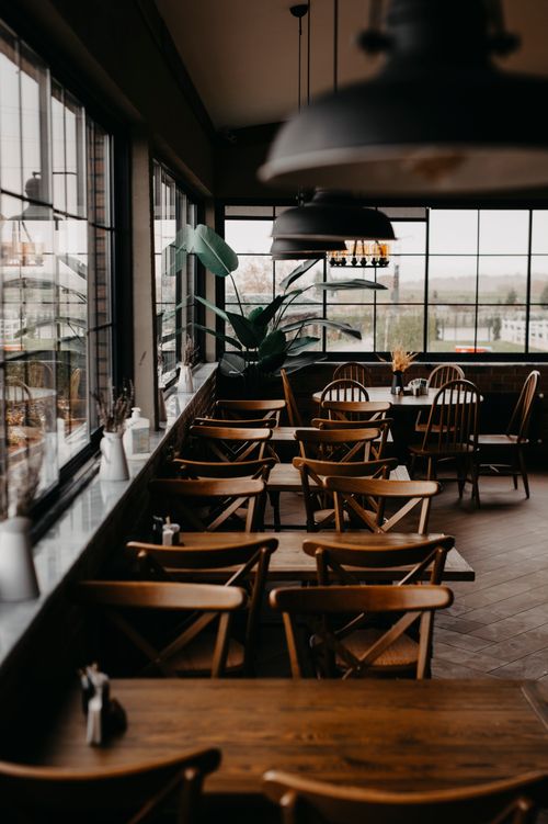 Free Warm restaurant setting with wooden tables and chairs, potted plants, and large windows. Stock Photo