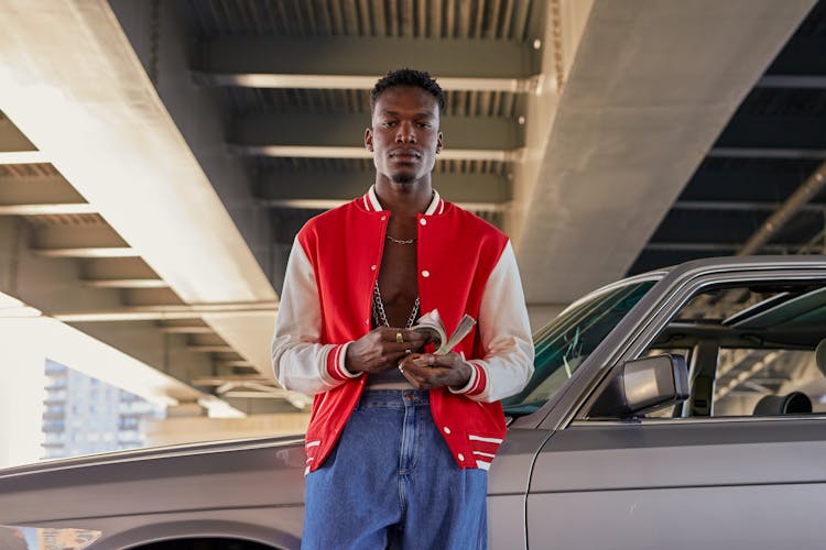 A Man In Red And White Jacket Leaning On The Car While Holding Money