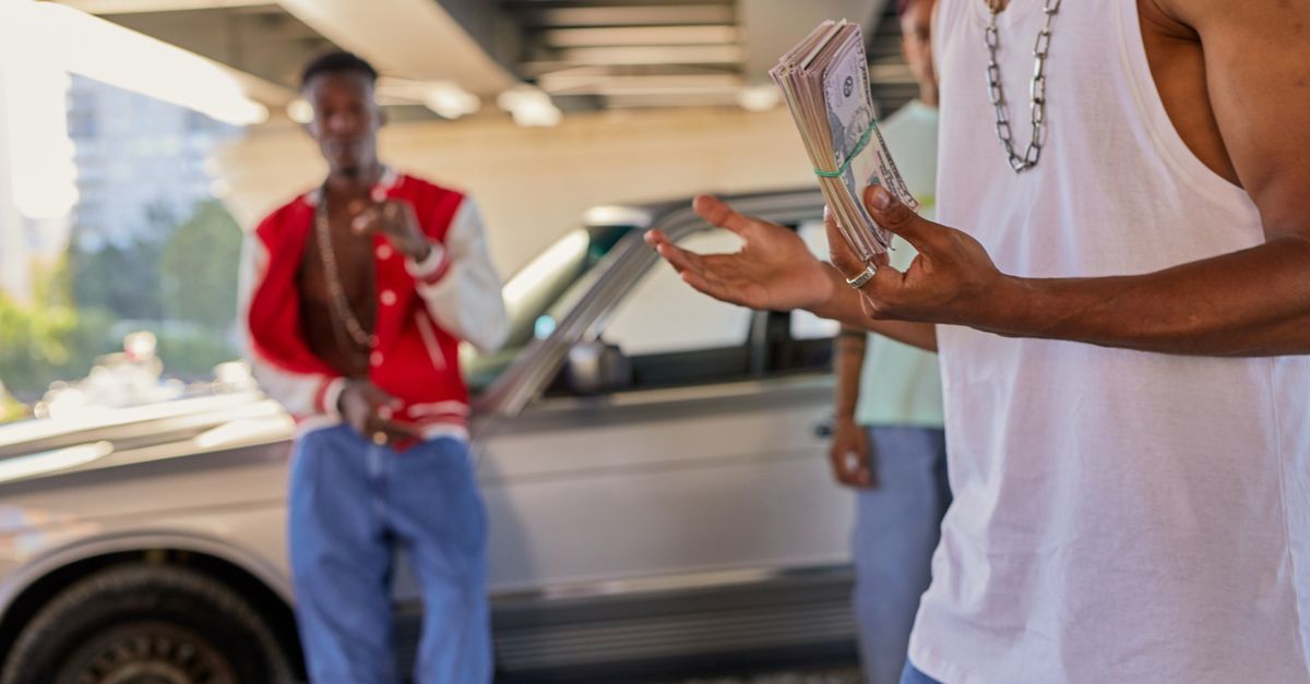 Young men handling cash bundles under a bridge near a parked car in daylight.