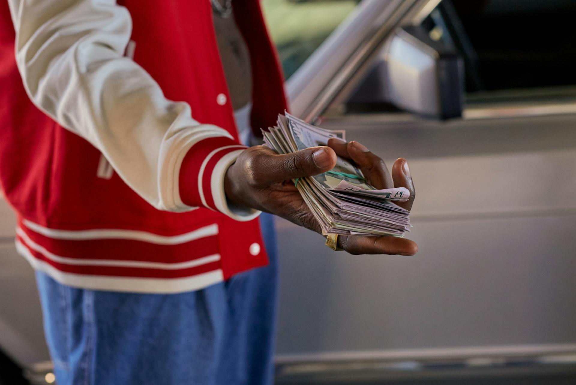 Close-up of a person offering a stack of cash in front of a car, symbolizes financial transaction.