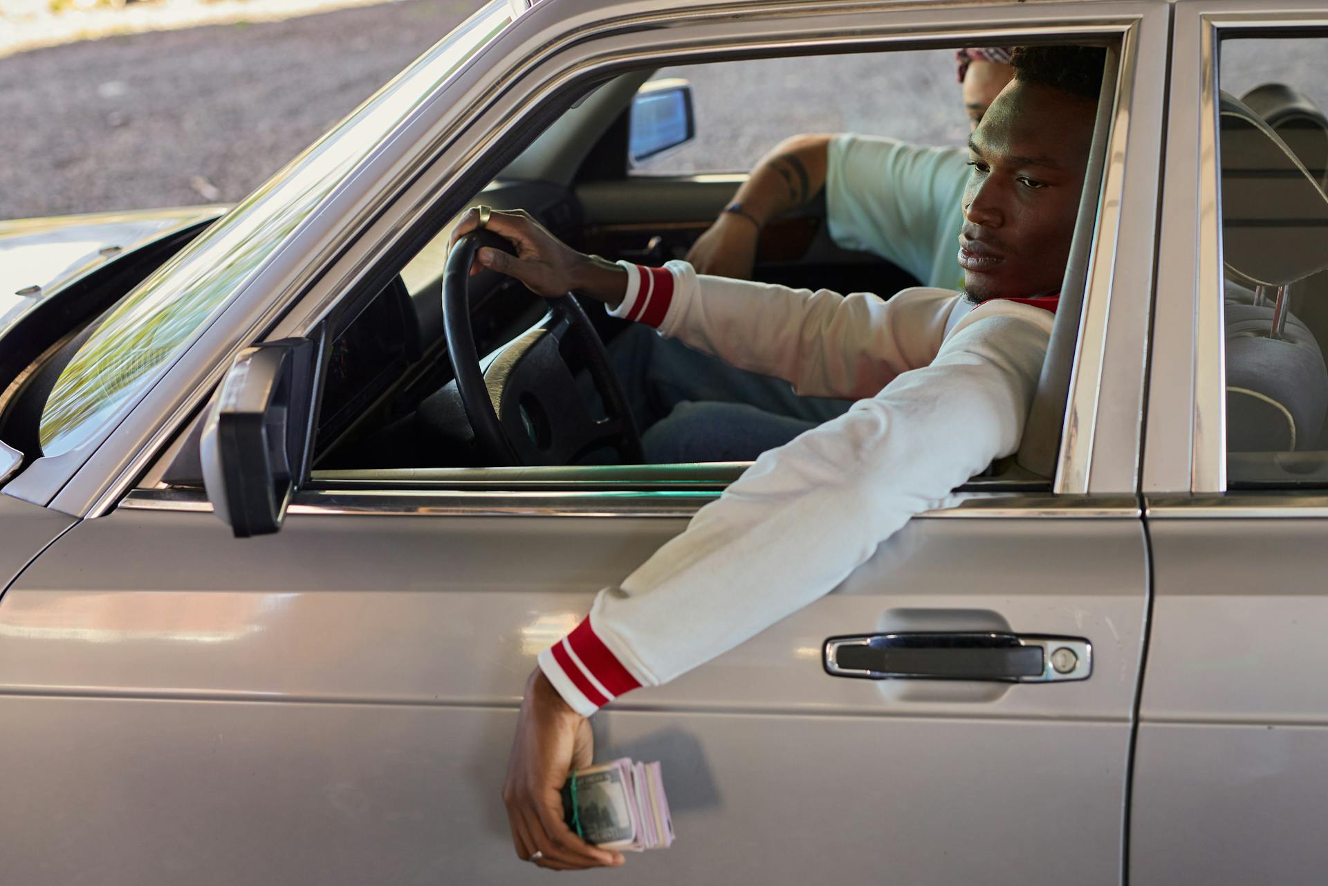 A young adult man sits in a vintage car, casually holding a wad of cash through the window.