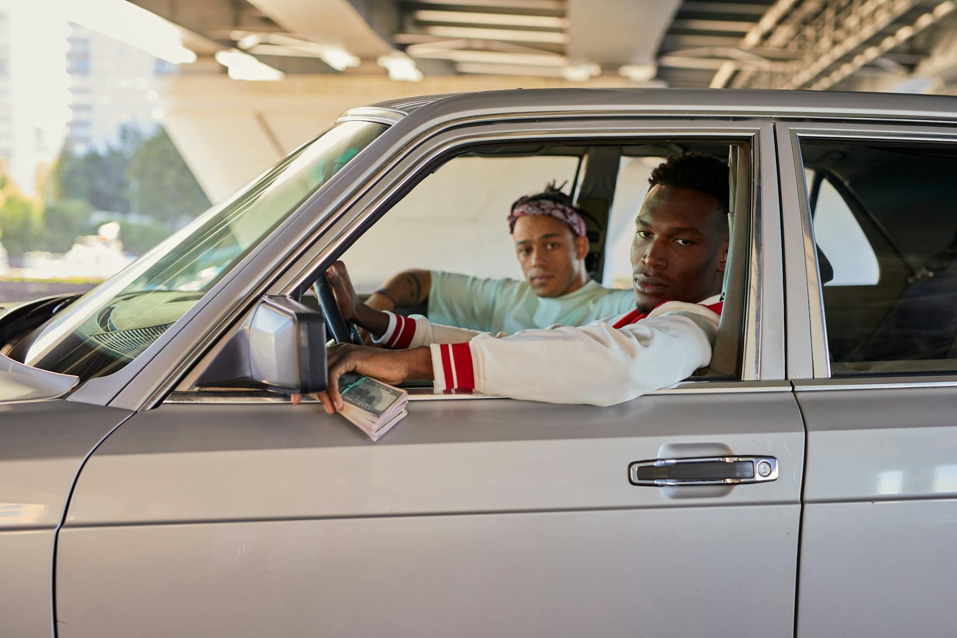 Two men seated in a car under a bridge, holding cash with a serious expression.