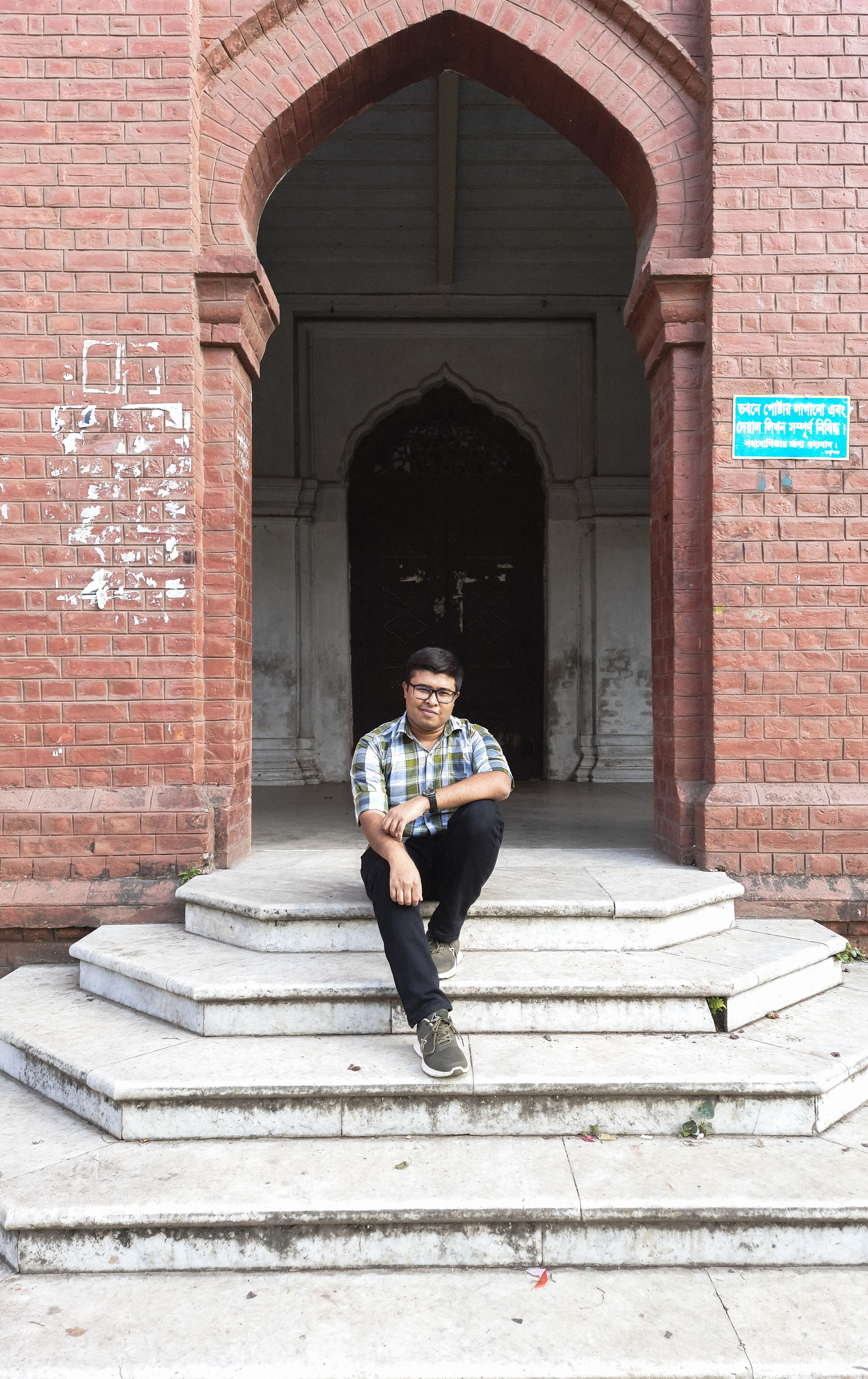 Asian man sitting on white concrete steps in front of a brick arch building in Dhaka, Bangladesh.