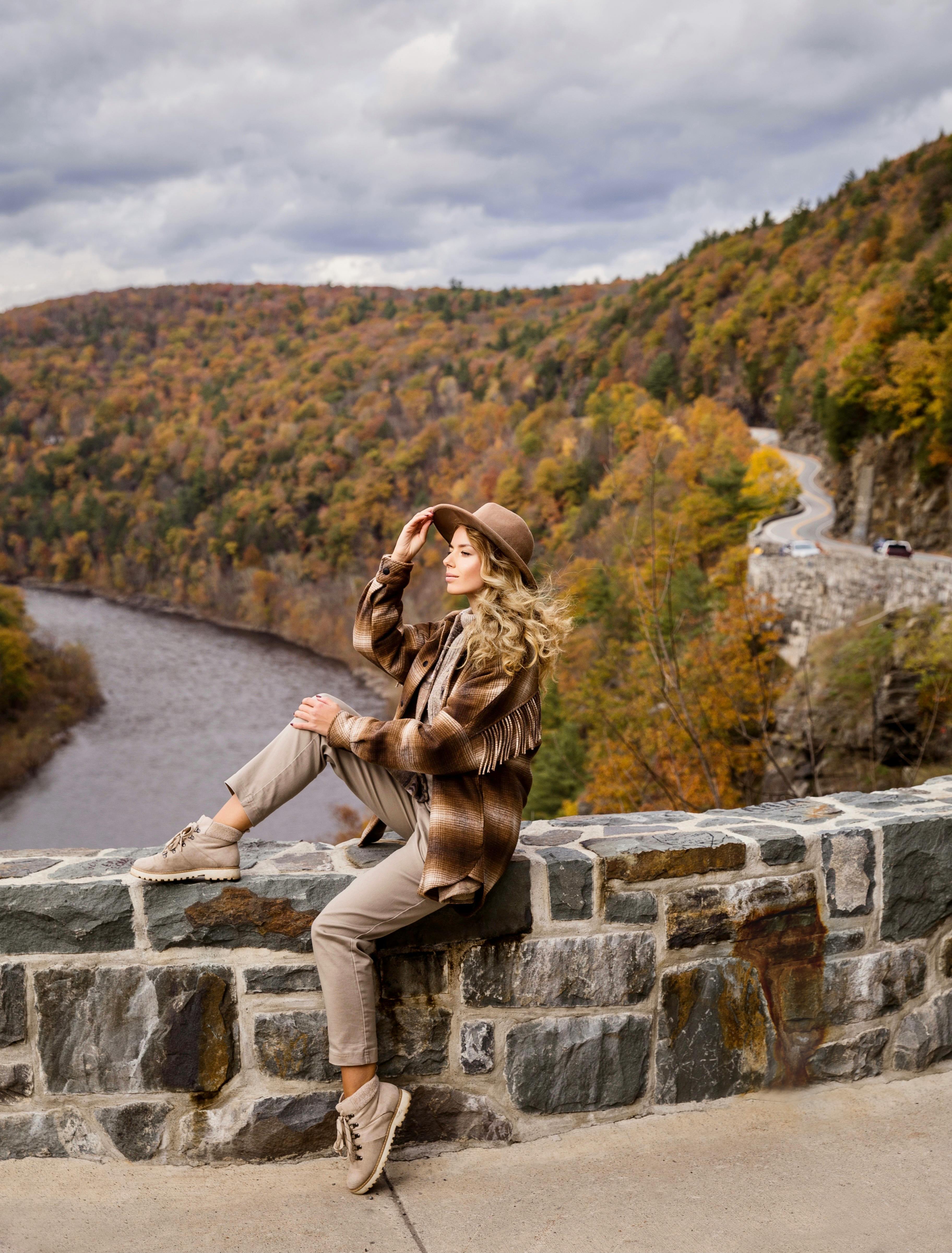 blond woman in tourist outfit sitting on stony bridge over mountain river