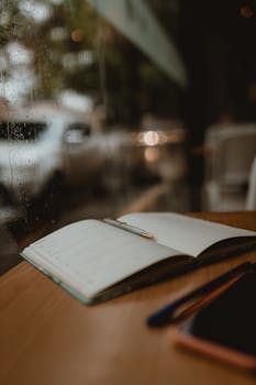 A cozy close-up of an open notebook and pen on a wooden table by a rainy window.