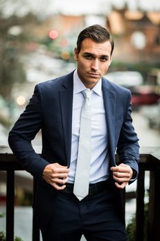 A confident businessman adjusts his suit jacket outdoors with a defocused city background.