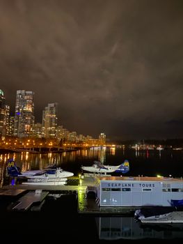 Illuminated city skyline with seaplanes docked along a waterfront under an overcast night sky.