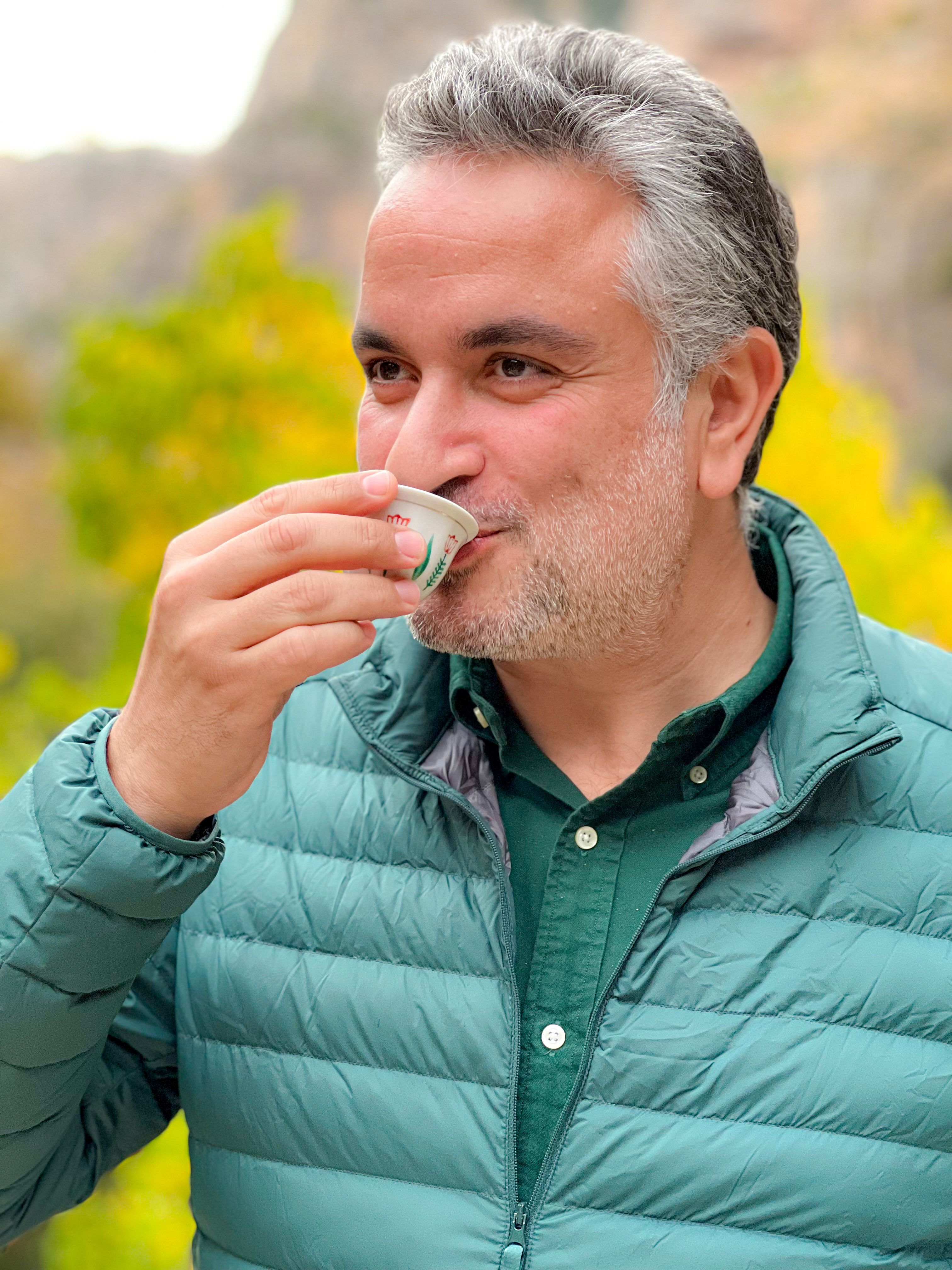 Man with gray hair wearing green jacket, savoring a hot drink outdoors in a colorful setting.