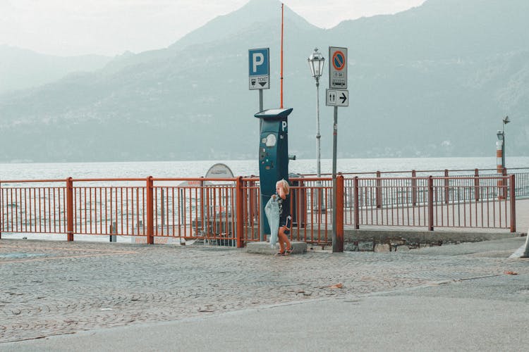 Child Standing On Gray Stone Pavement Beside A Parking Ticket Booth